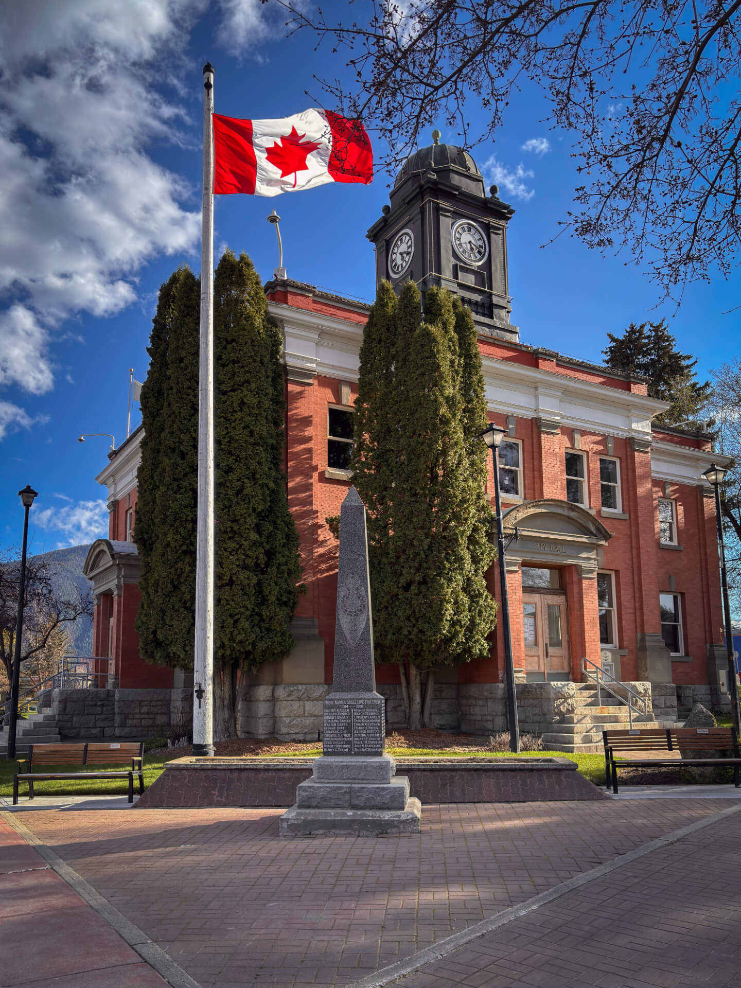 Grand Forks City Hall, a red-brick building with a black clock tower, with the Canadian flag flying on a tall pole and the stone cenotaph on the plaza in front